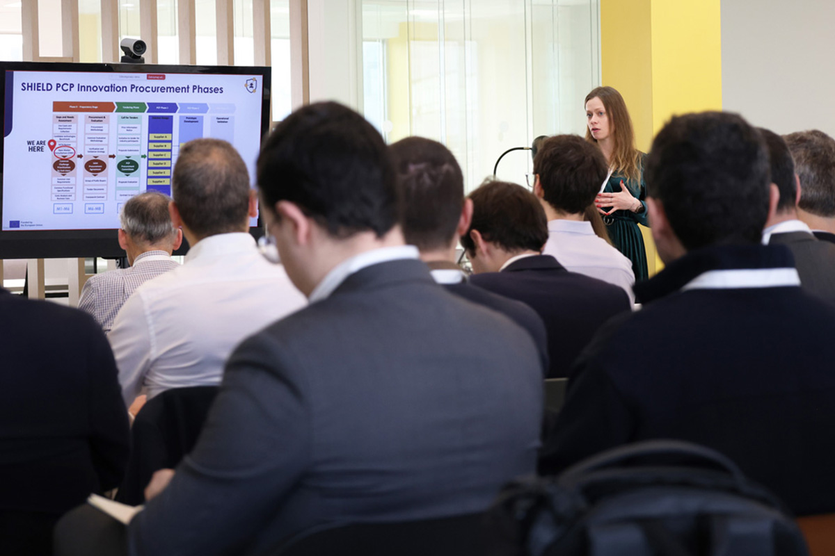 A speaker addresses an audience in a conference room beside a screen displaying the SHIELD PCP Innovation Procurement Phases.
